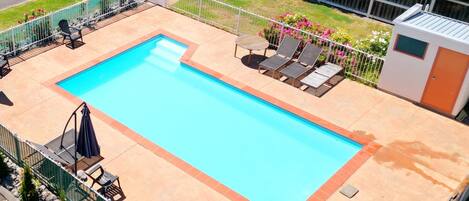 A rectangular swimming pool with a tiled deck, surrounded by a white fence. There are lounge chairs and umbrellas along the poolside.