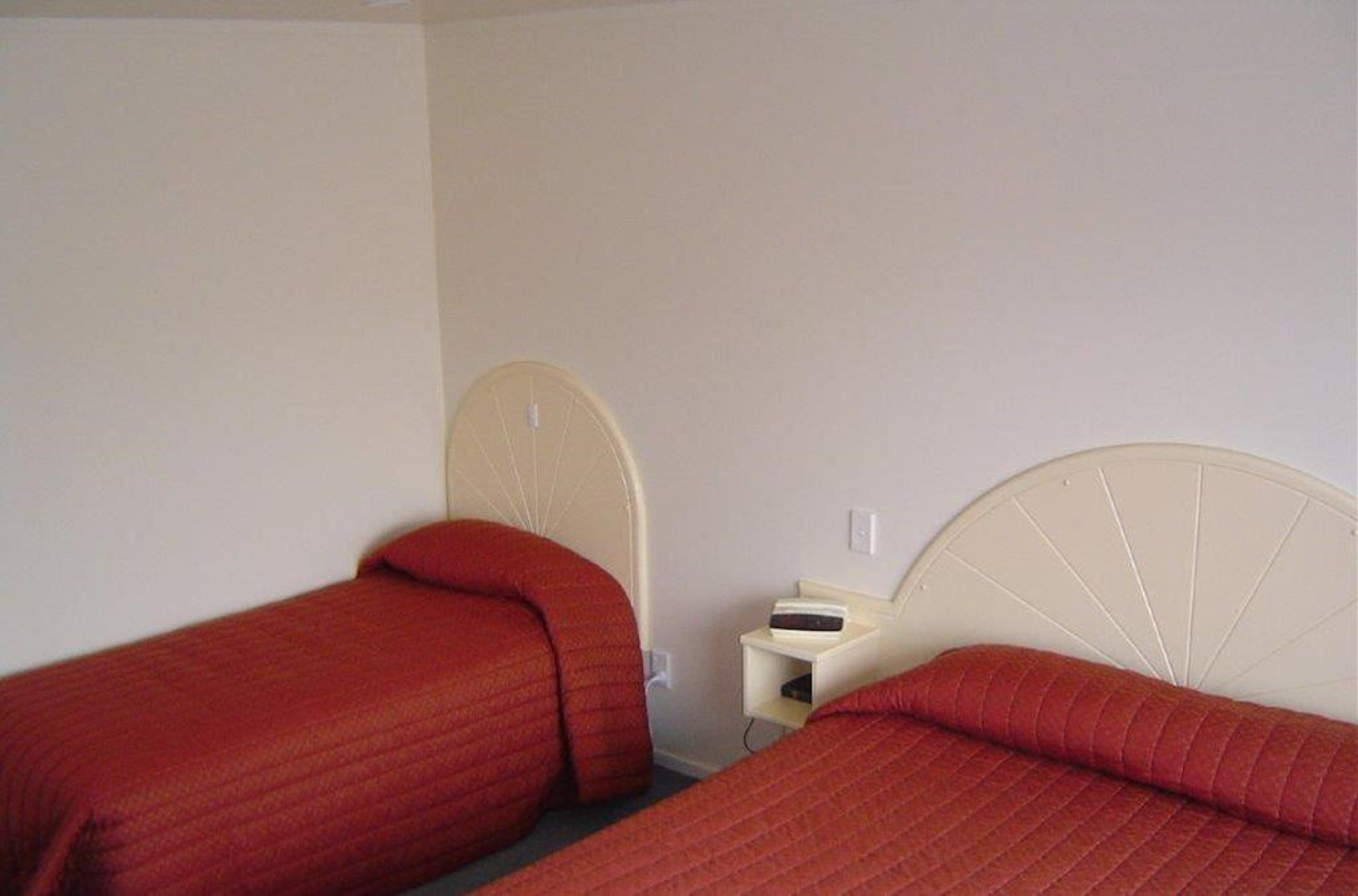A room with two single beds, each with a headboard and a red quilted bedspread. There is a small bedside table with a lamp and a power outlet.