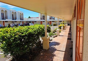 A covered walkway with a row of rooms, a parking area, and greenery. - Valdez Motor Lodge (Hastings)