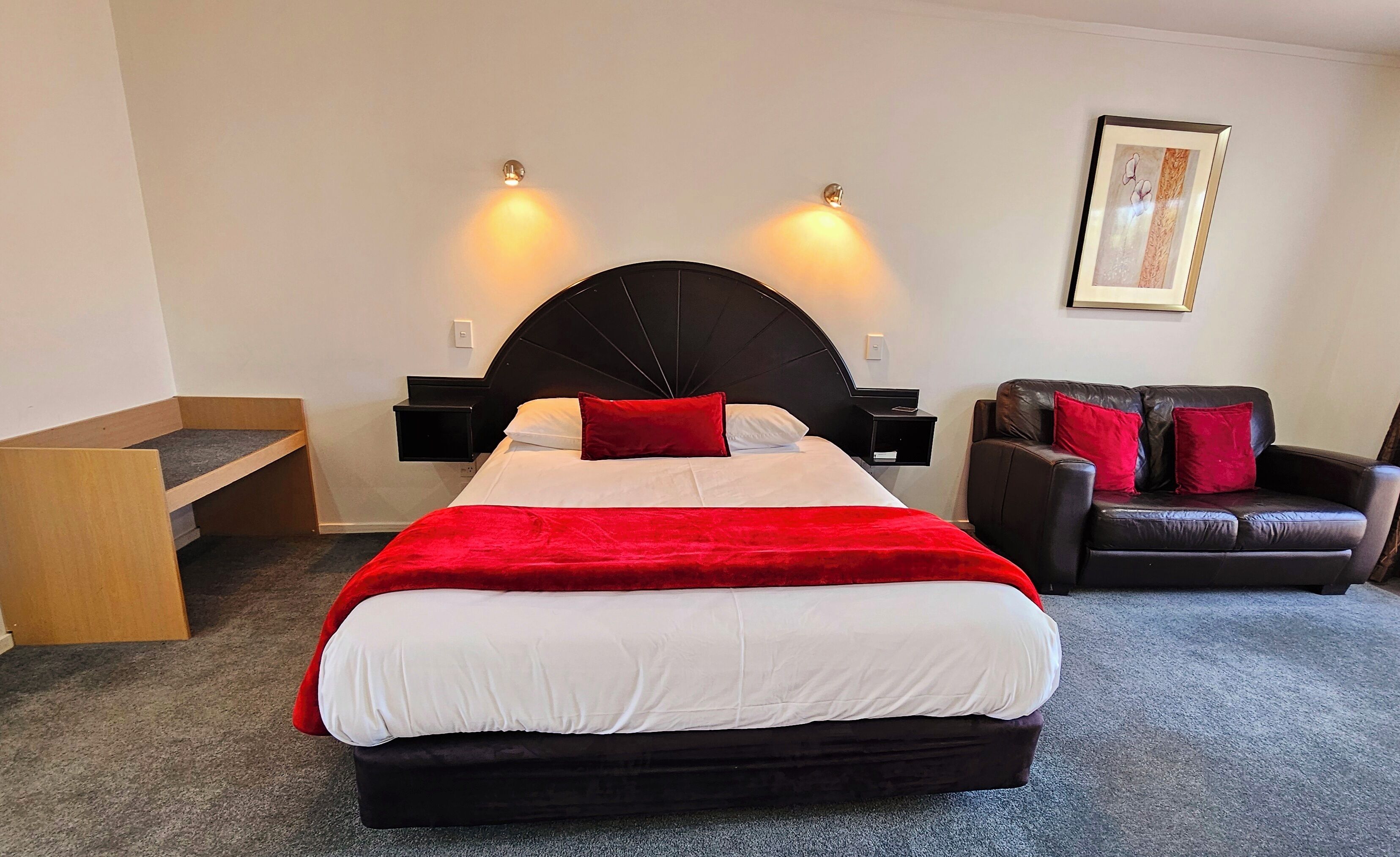 A hotel room with a large bed, a black headboard, two bedside tables, a red throw blanket, a black leather sofa with red cushions, and a framed picture on the wall.