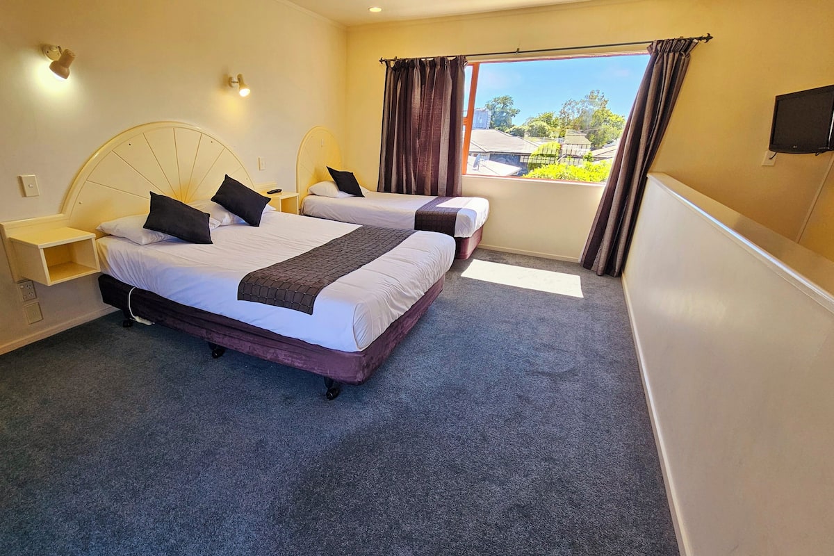 A hotel room with two beds, a large window with curtains, a flat-screen TV, and a small bedside table with a lamp.