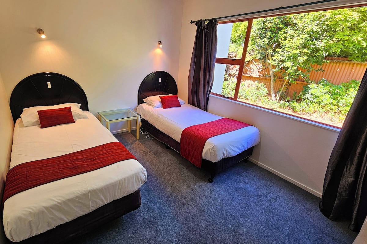 A bedroom with two single beds, a small table, a window with curtains, and a view of greenery outside.