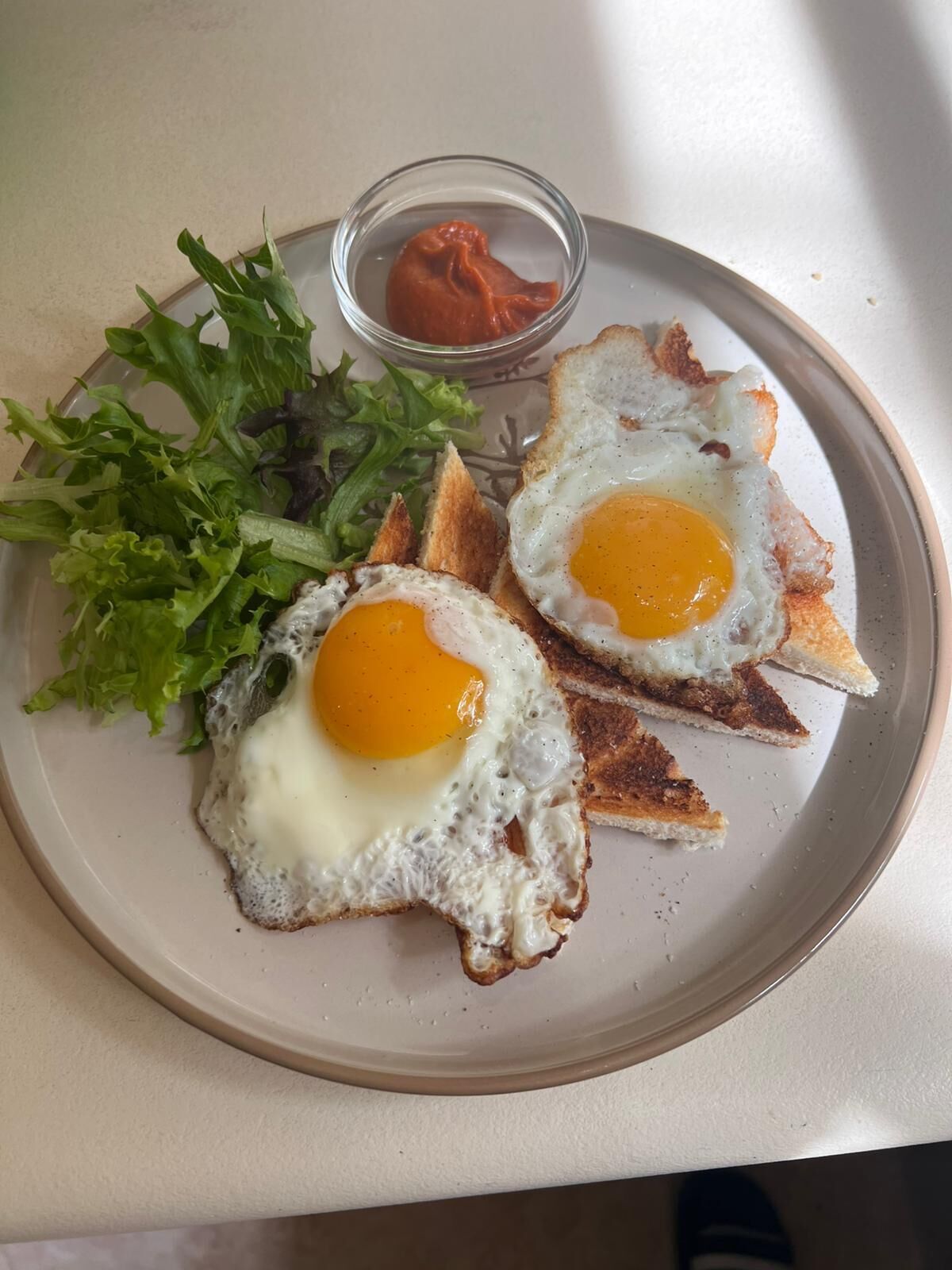 Un plato con dos huevos fritos sobre tostada, una ensalada y un recipiente pequeño con salsa.