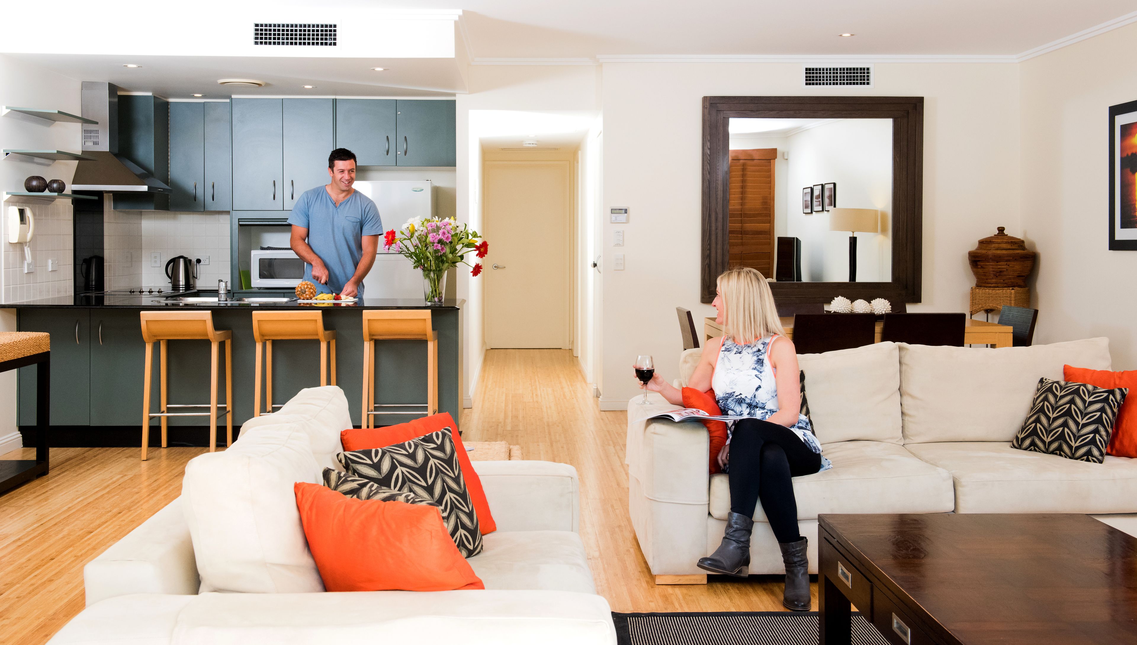 A modern kitchen with dark cabinets and a white countertop, a living room with a white sofa and orange pillows, and a dining area with bar stools.