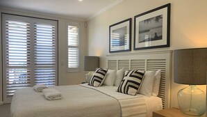 Un dormitorio con cama, almohadas, una mesita de noche, una lámpara y una ventana con persianas.