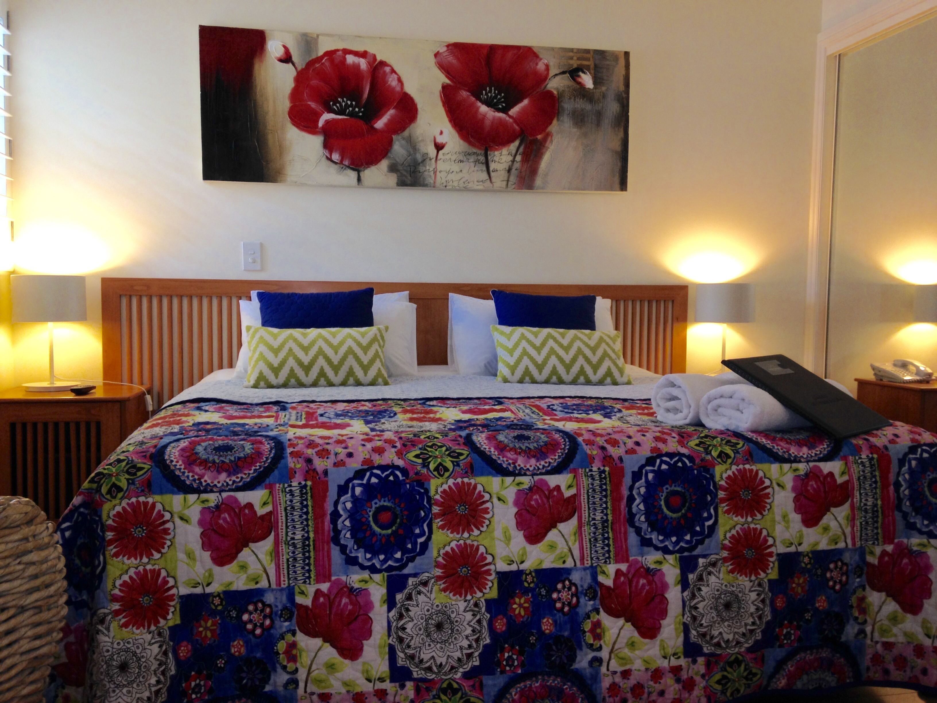 Quarto de hotel com cabeceira de madeira, duas almofadas, colcha colorida e pintura de flores vermelhas na parede.