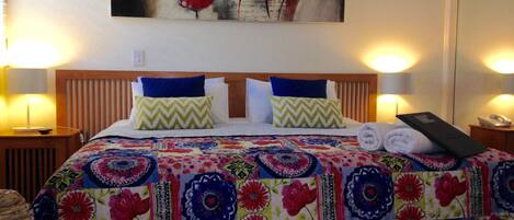 Quarto de hotel com cabeceira de madeira, duas almofadas, colcha colorida e pintura de flores vermelhas na parede.