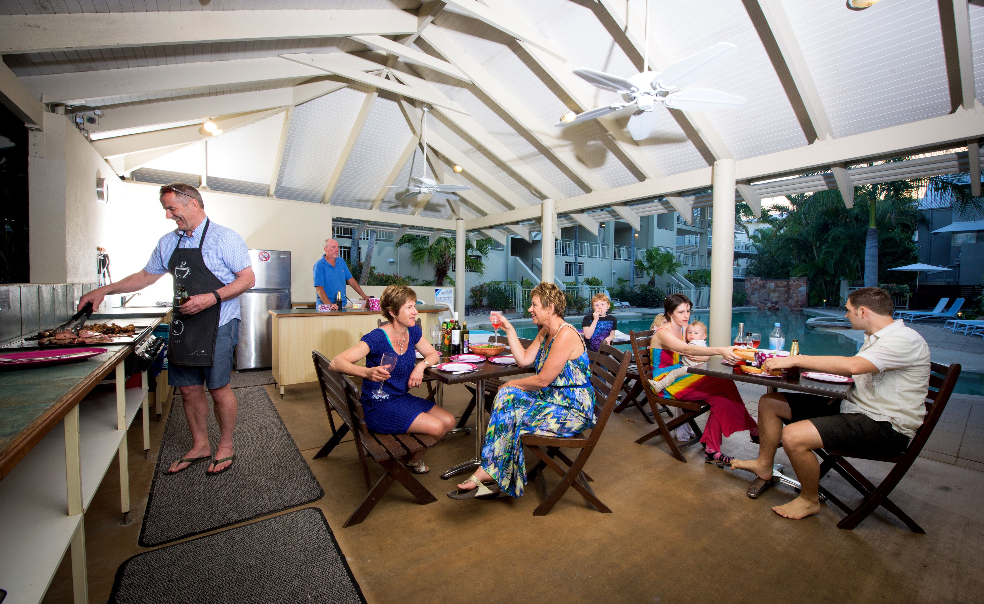 An outdoor dining area with a pool view, covered by a white roof with ceiling fans, and a bar area with a man grilling.