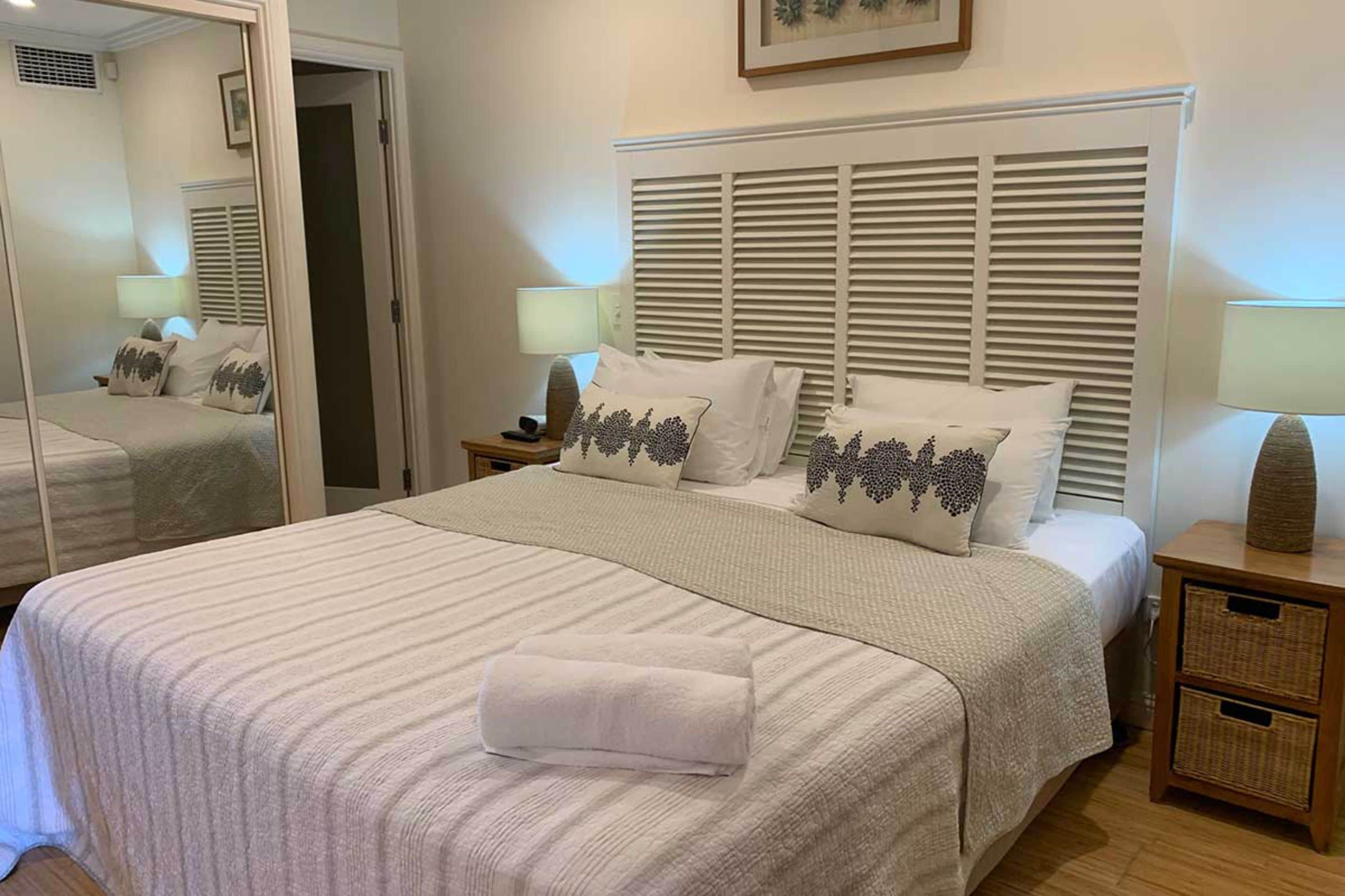 Un lit bien fait, recouvert d’une couvre-lit blanche et agrémenté d’oreillers assortis, une table de chevet avec une lampe et un miroir qui reflète la pièce.