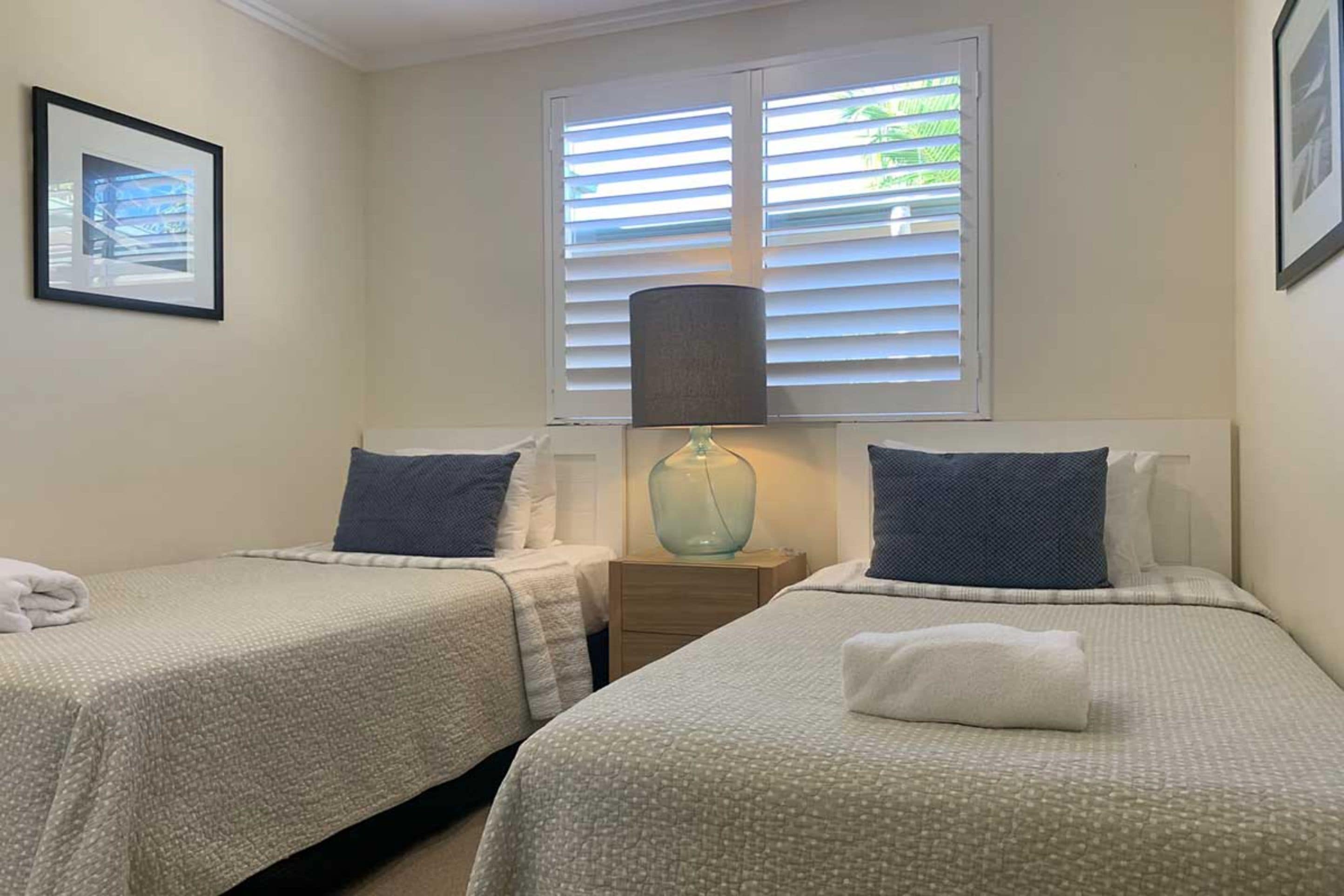 Une chambre avec deux lits, une table de chevet, une lampe et une fenêtre avec des stores.