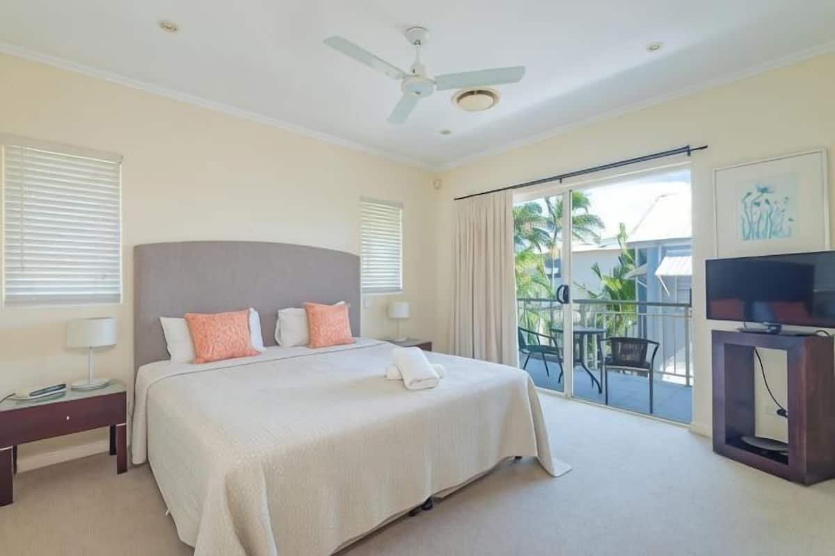 A hotel room with a bed, a ceiling fan, a TV stand, and a balcony with a view.