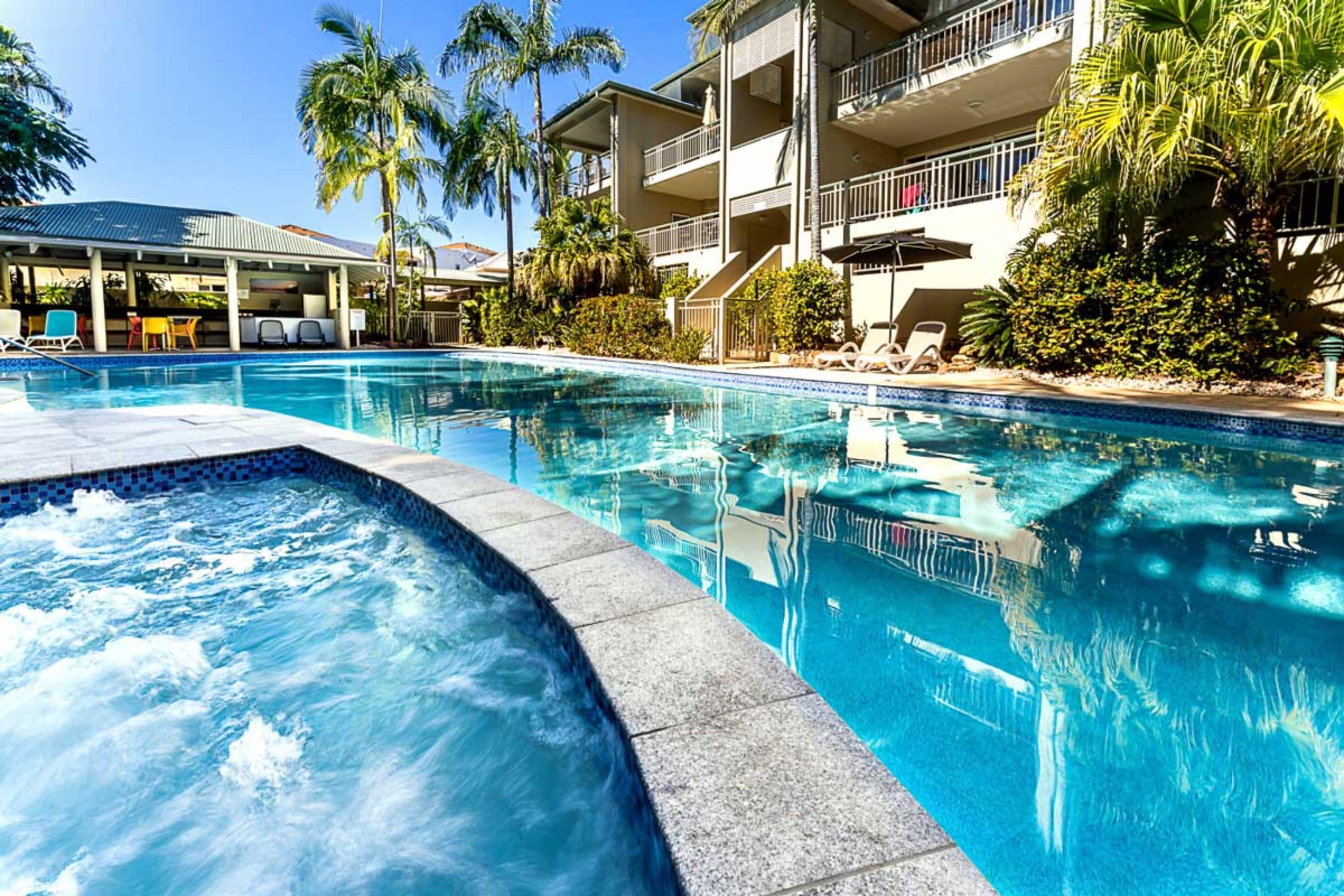 Una piscina con sillones de descanso y palmeras, rodeada por un edificio de varios pisos con balcones.