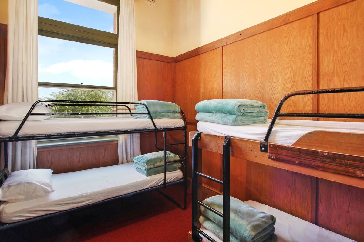 A room with two bunk beds, wooden walls, a window with curtains, and a view of trees outside.