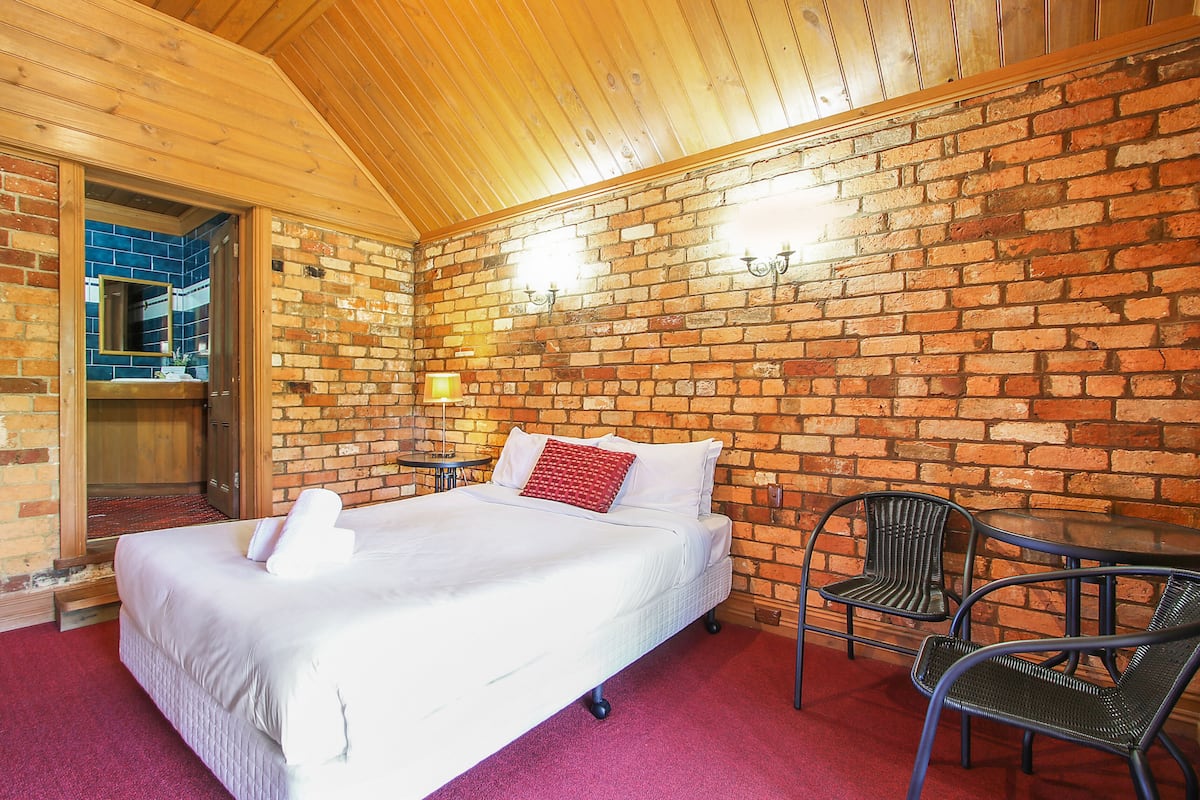 A room with a brick wall, a bed with white bedding, a red pillow, and two chairs.
