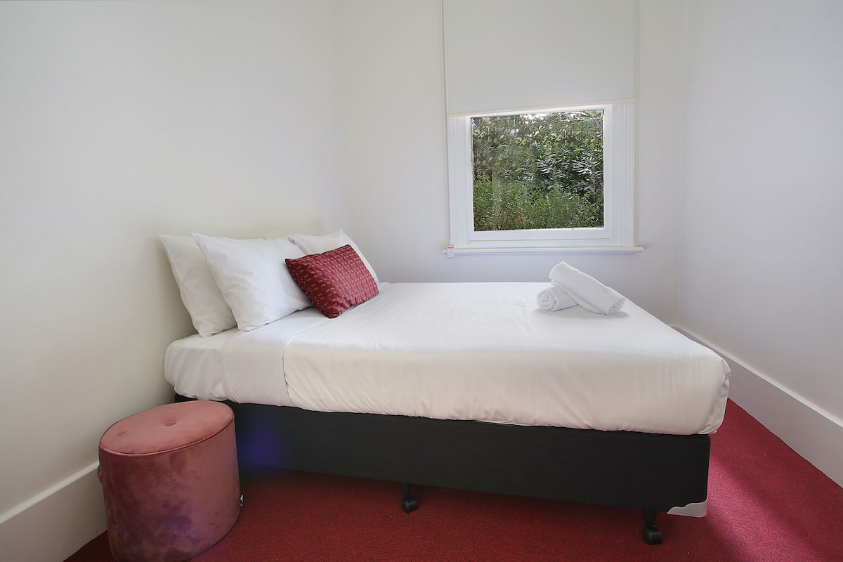 A neatly made bed with white bedding and a red pillow, a small round ottoman, and a window with a view of greenery.