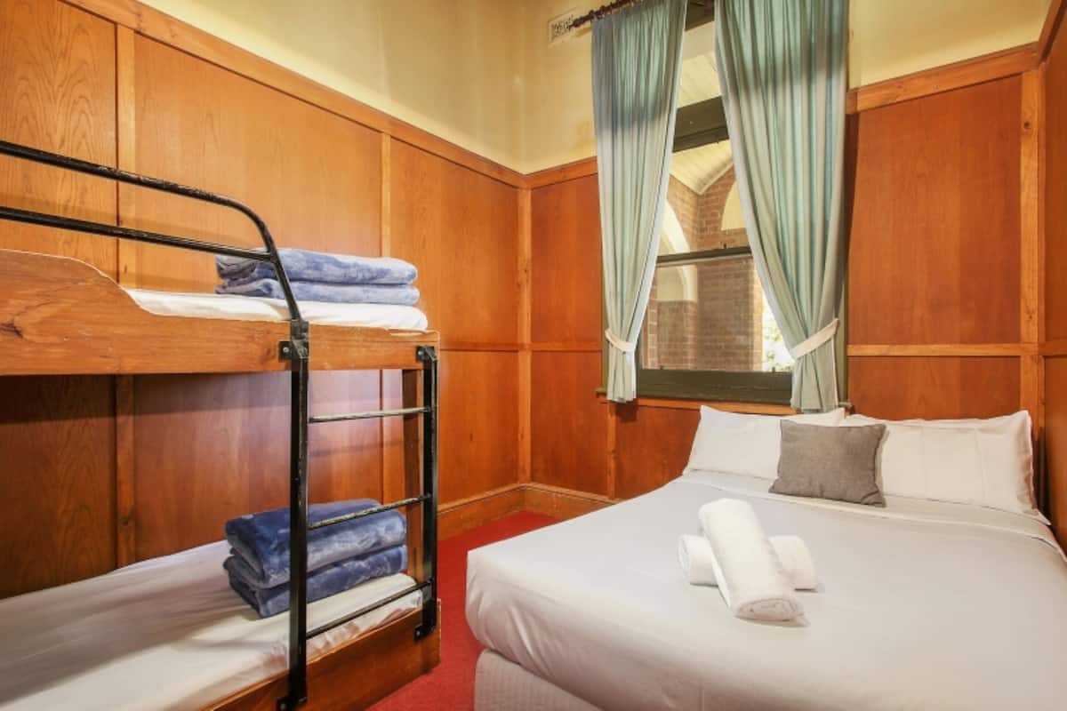 A room with a bunk bed, a single bed, wooden paneling, a window with curtains, and a brick wall visible through the window.