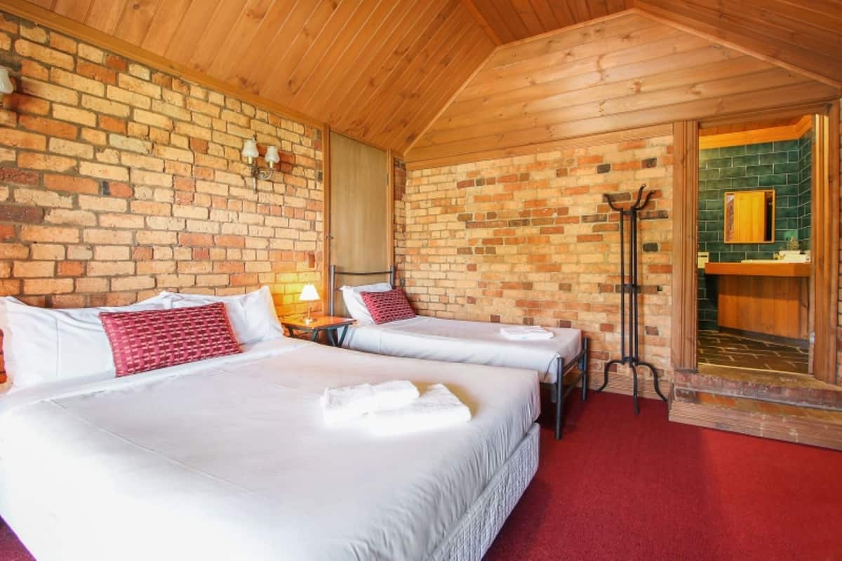 A room with two beds, wooden ceiling, brick walls, and a bathroom with a green-tiled shower area.