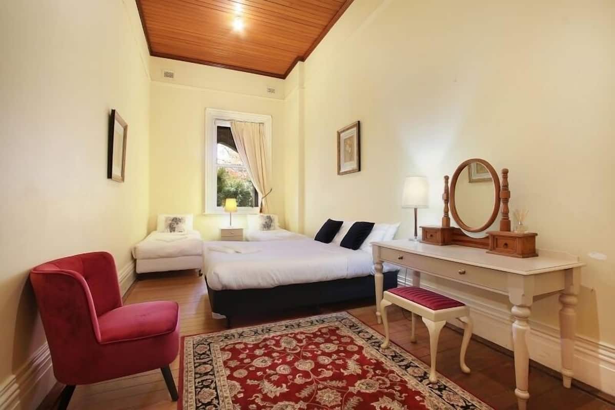 A bedroom with a bed, a red chair, a white dresser with a mirror, and a red and black patterned rug.