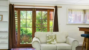 A room with a white sofa, a wooden floor, a sliding glass door with a view of greenery, and a small table with a chair. - The Priory Beechworth (Beechworth)