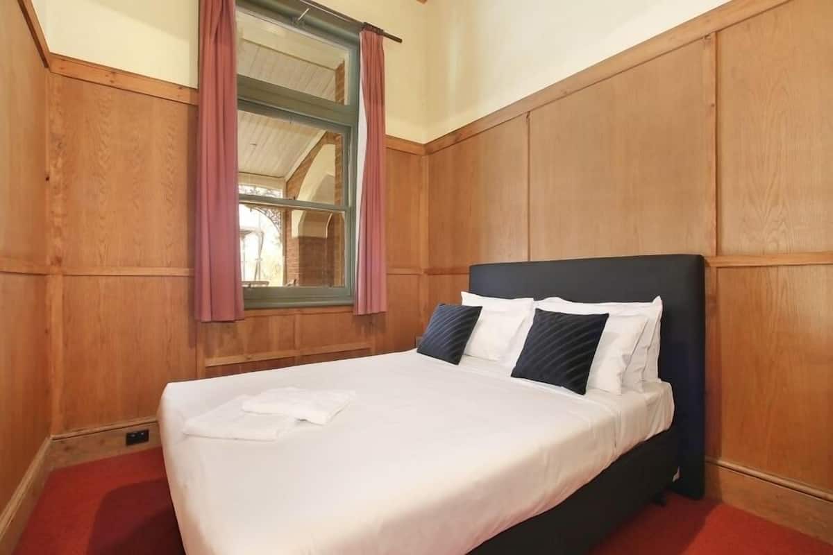 A neatly made bed with white linens and a folded towel, wooden paneling on the walls, and a window with pink curtains.