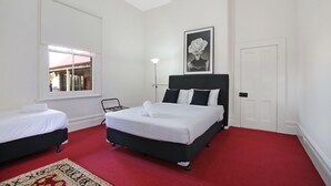 A hotel room with two beds, a red carpet, a white wall, a framed picture, and a window with a view. - The Priory Beechworth (Beechworth)