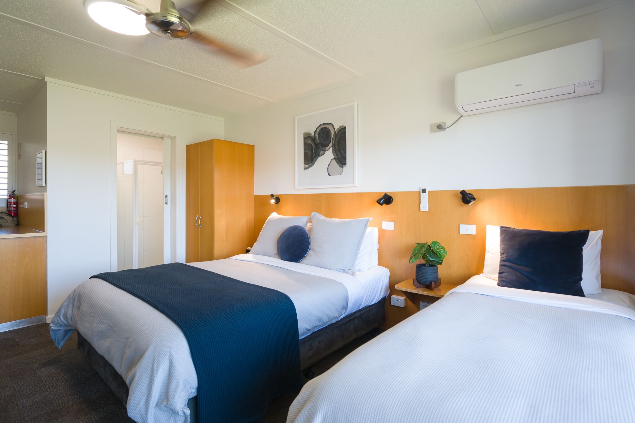 Une chambre d’hôtel avec deux lits, un ventilateur de plafond, un appareil de climatisation, un tableau encadré, des lampes de chevet et une petite plante en pot.