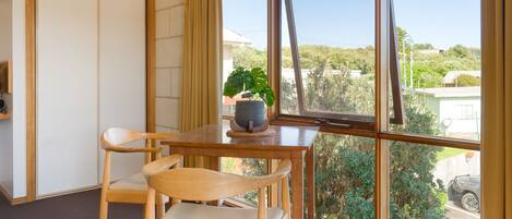 Una habitación con una mesa de madera y dos sillas, una planta en maceta y vistas a una zona residencial a través de amplias ventanas.