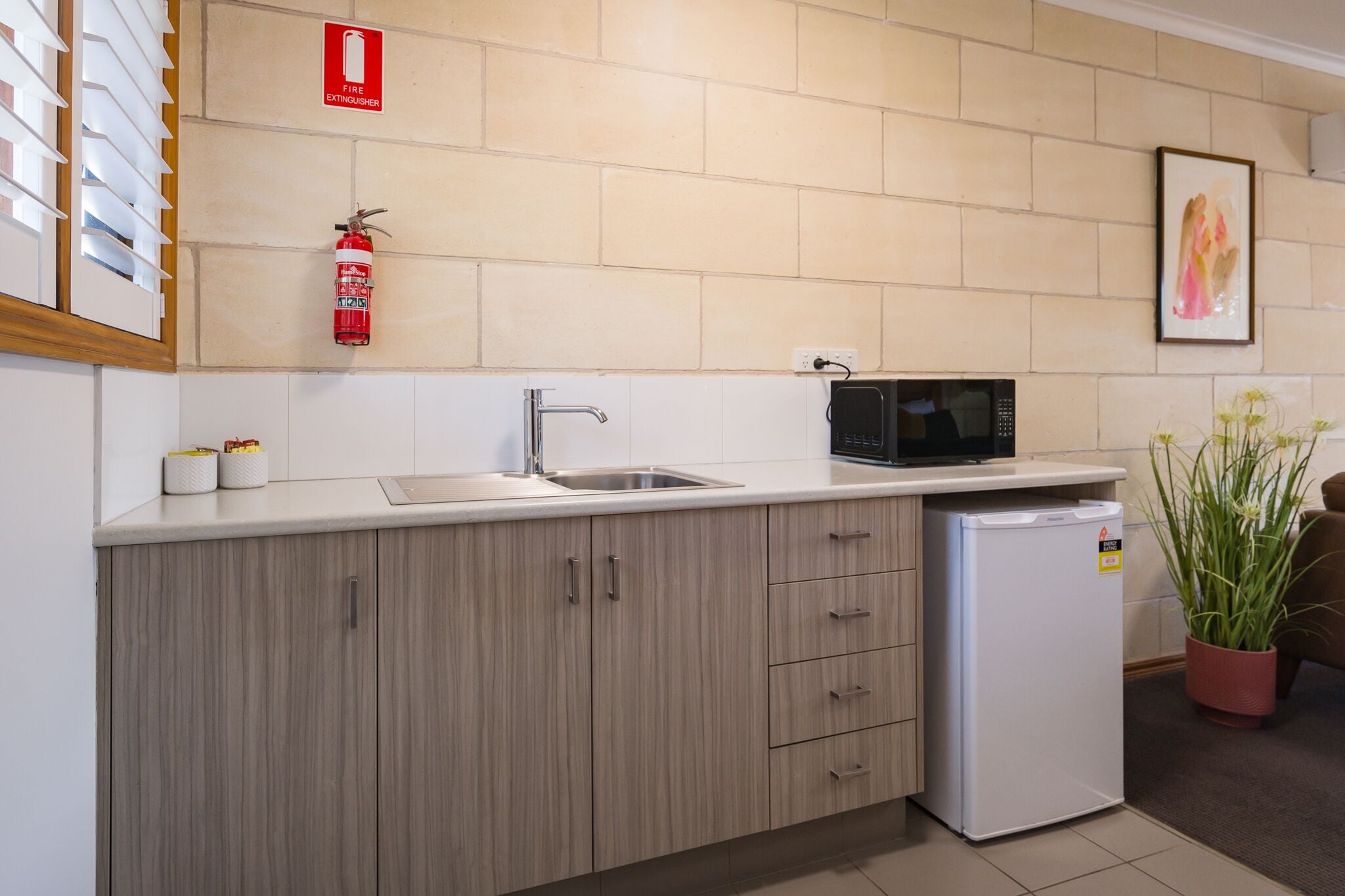 Une cuisine moderne équipée d’un évier, d’un four à micro-ondes et d’un petit réfrigérateur. Un extincteur est fixé au mur, et un tableau encadré est accroché au-dessus de l’évier.