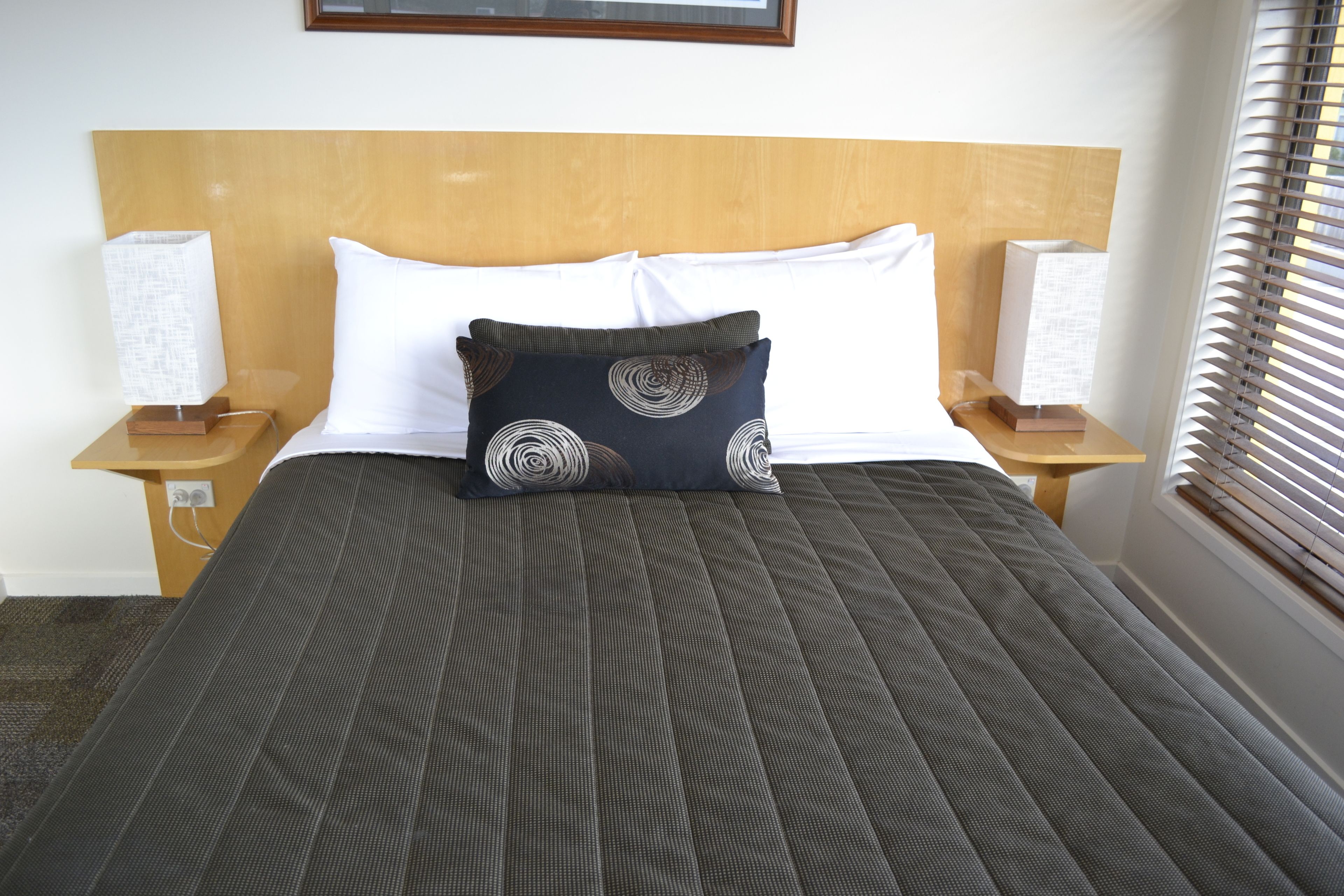 A neatly made bed with a dark quilt, two white pillows, and a decorative pillow with circular patterns. There are two bedside tables with lamps and a window with blinds.