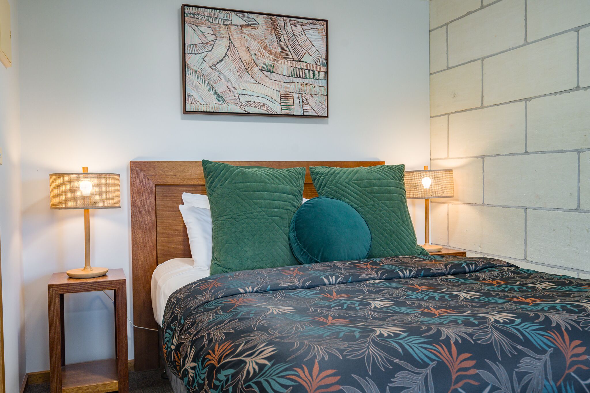 A neatly made bed with a patterned bedspread, two green pillows, and a wooden headboard. There is a wall-mounted abstract art piece above the bed and a brick-patterned wall to the right.