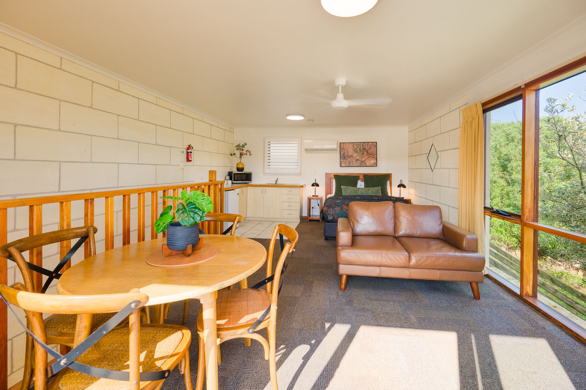 A cozy living area with a wooden dining table, leather sofa, and a view of greenery through large windows.