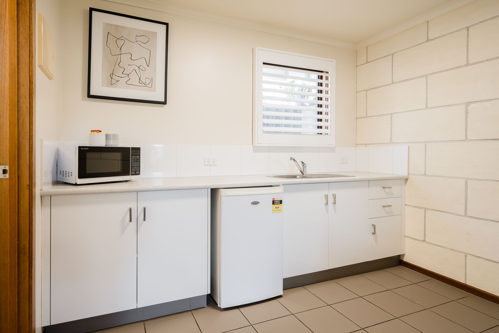 A compact kitchen with white cabinets, a microwave, a refrigerator, a sink, and a window with blinds.