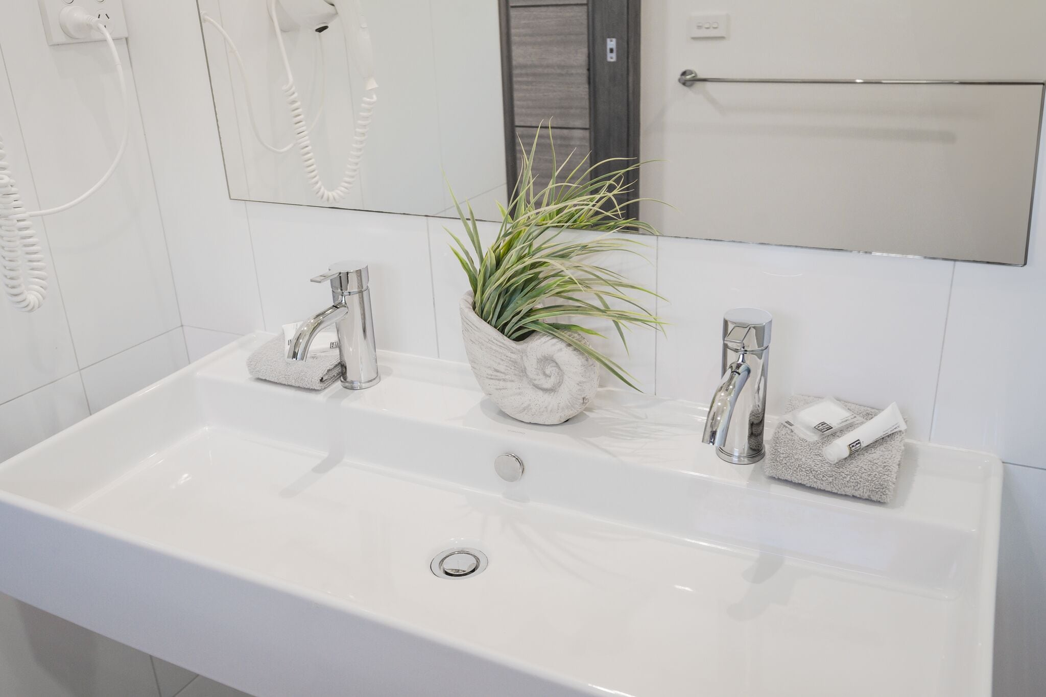 A white bathroom sink with a plant in a white pot, two towel holders, and a mirror above.