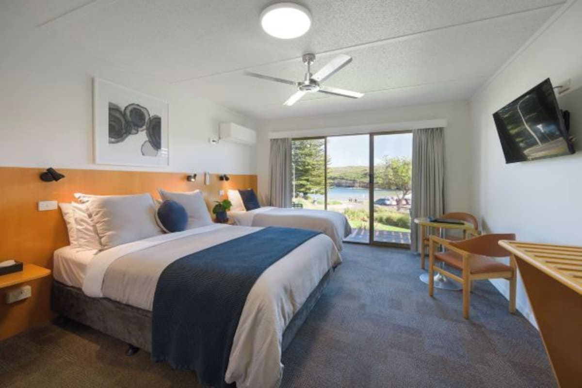 A hotel room with two beds, a desk, a chair, a ceiling fan, and a large window with a view of the outdoors.