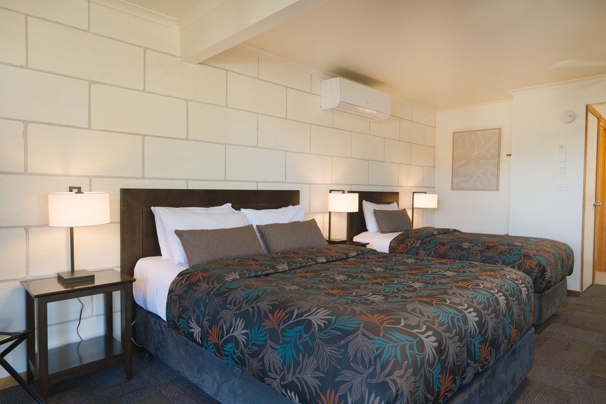 A hotel room with two beds, a nightstand, a lamp, and a wall-mounted air conditioner.