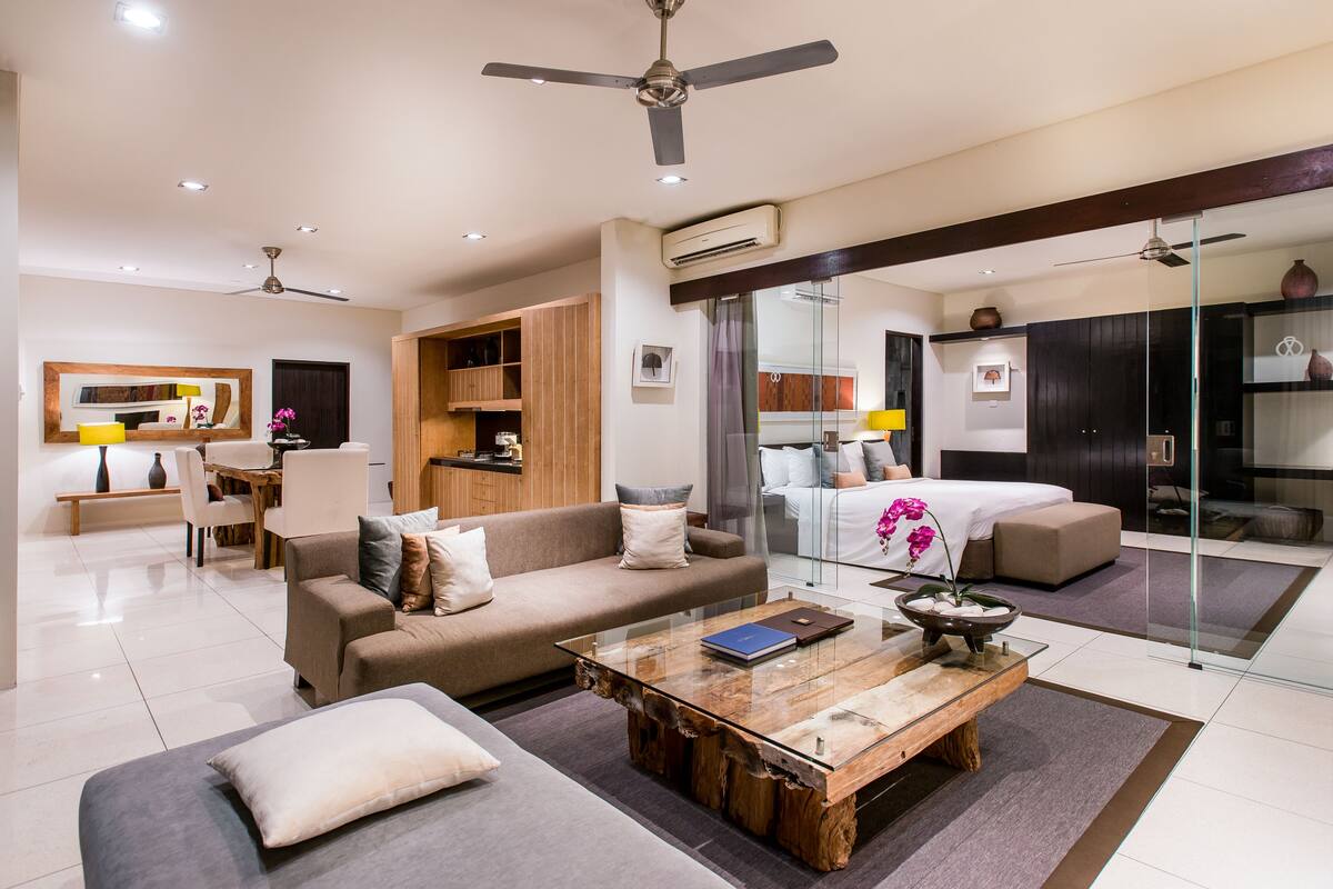 A modern living room with a sofa, coffee table, and a glass partition separating the living area from the bedroom.
