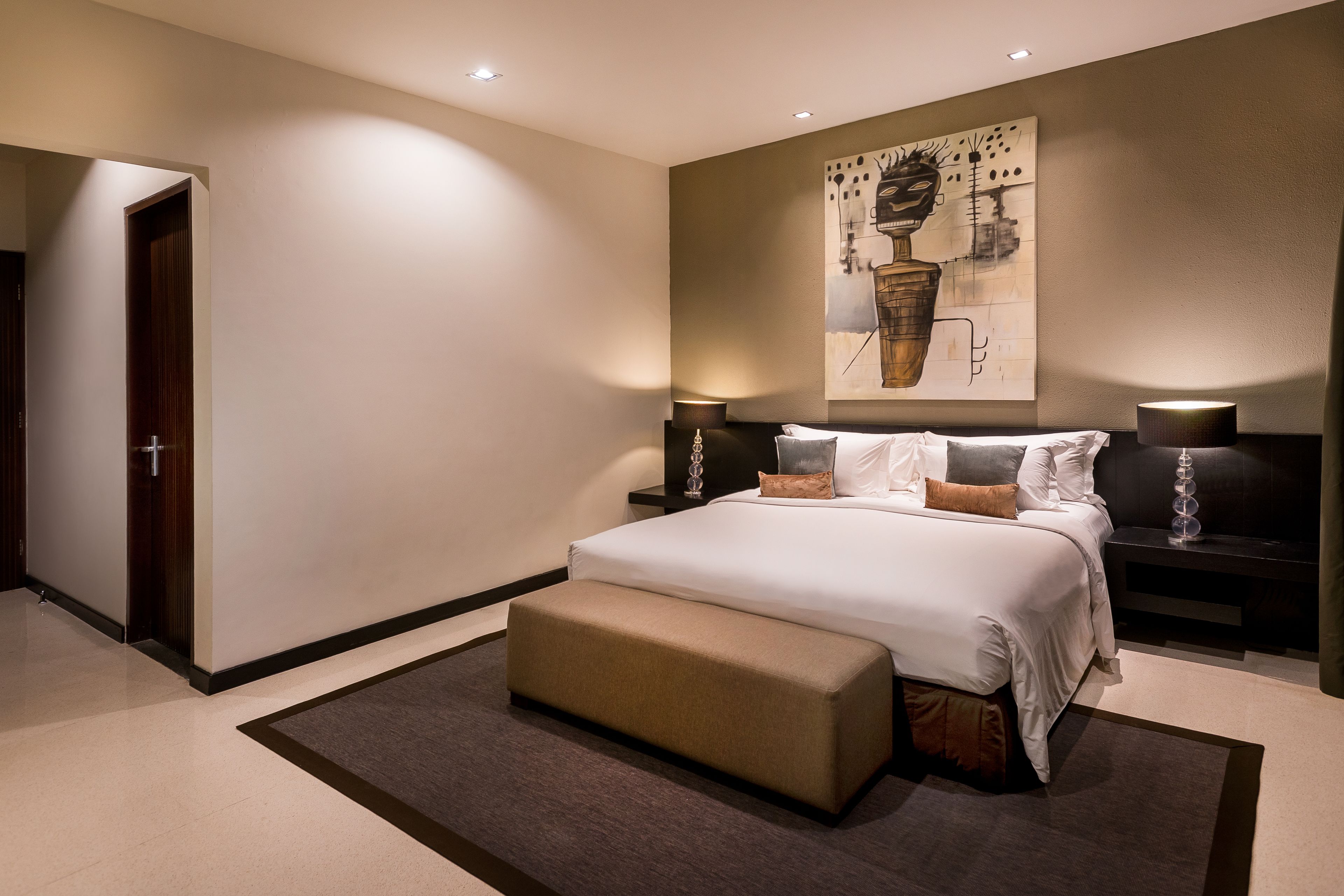 A modern hotel room with a large bed, a bench, a nightstand with a lamp, and a painting on the wall.