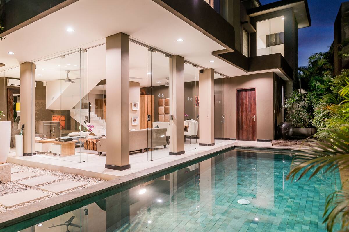 A modern poolside area with a glass-enclosed lounge, a staircase leading to upper levels, and a view of the pool.