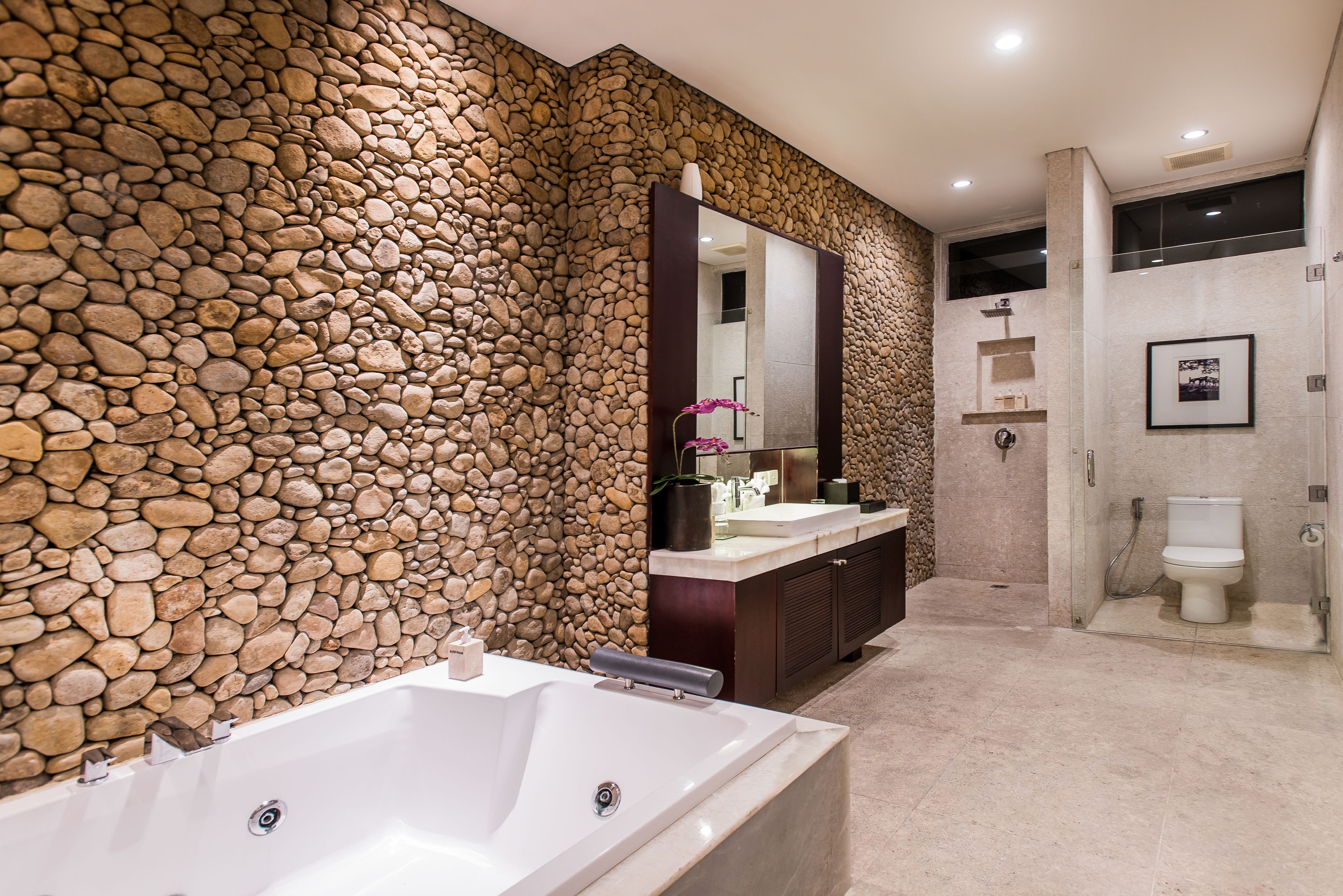 A modern bathroom with a stone wall, a large bathtub, a vanity with a mirror, and a toilet.