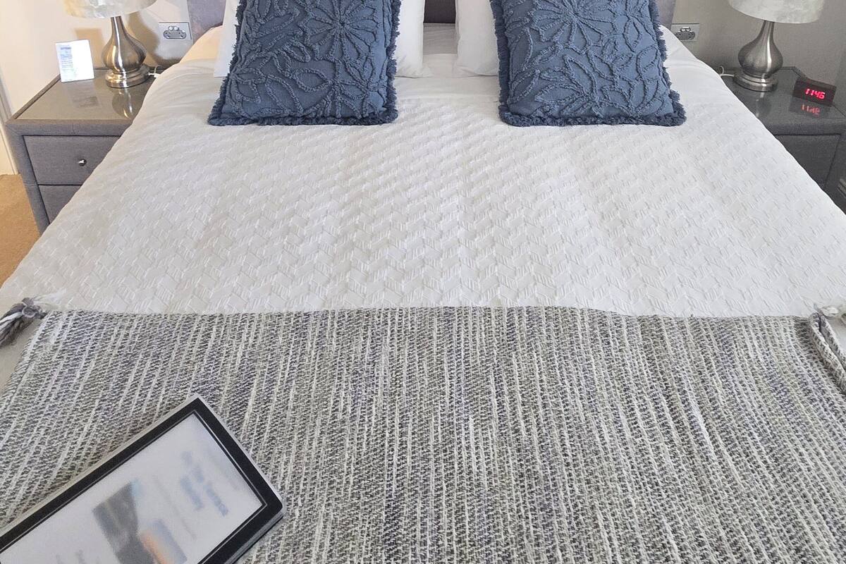 Un lit bien fait, avec une couette blanche, deux oreillers bleus et une tête de lit grise. Un tableau encadré est accroché au-dessus du lit, et une horloge numérique affiche l’heure.