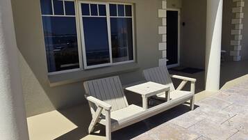 A patio with two white lounge chairs and a small table, a window with blinds, and a door to the right.