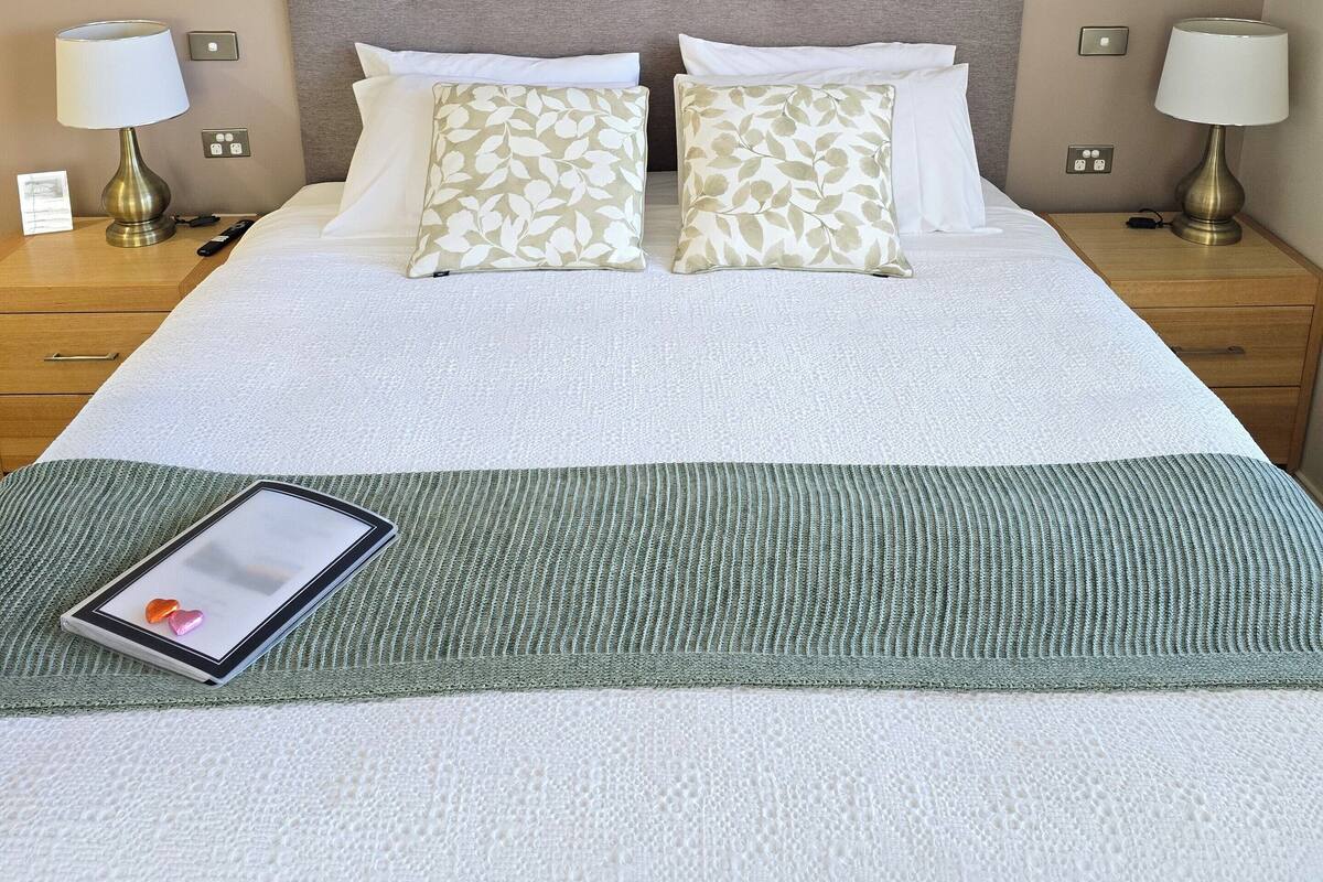 Un lit bien fait, recouvert d’un plaid vert, deux tables de chevet avec des lampes, une œuvre d’art accrochée au mur et une tablette posée sur le lit.