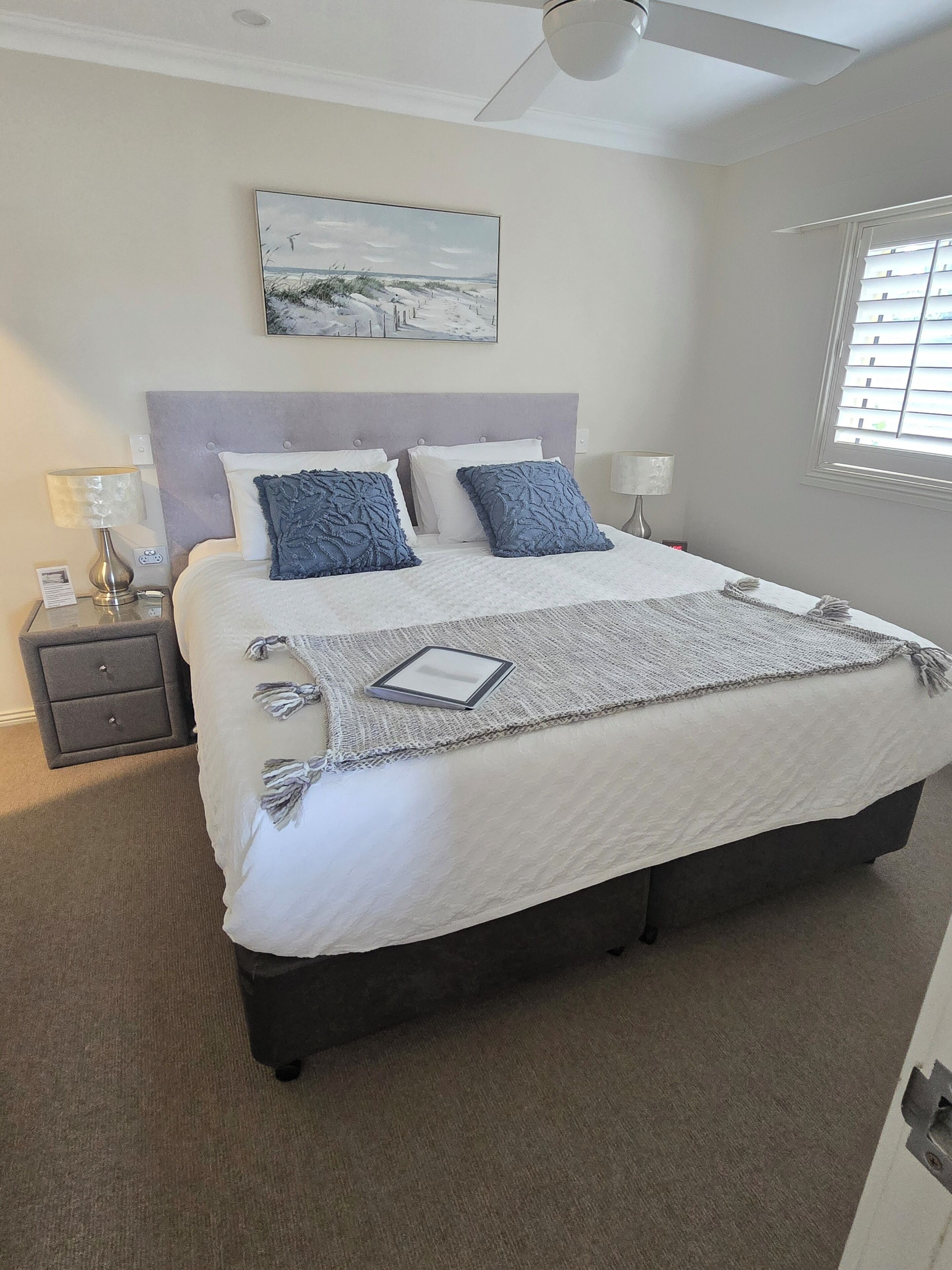 A bedroom with a large bed, a nightstand, a lamp, a ceiling fan, and a window with blinds.