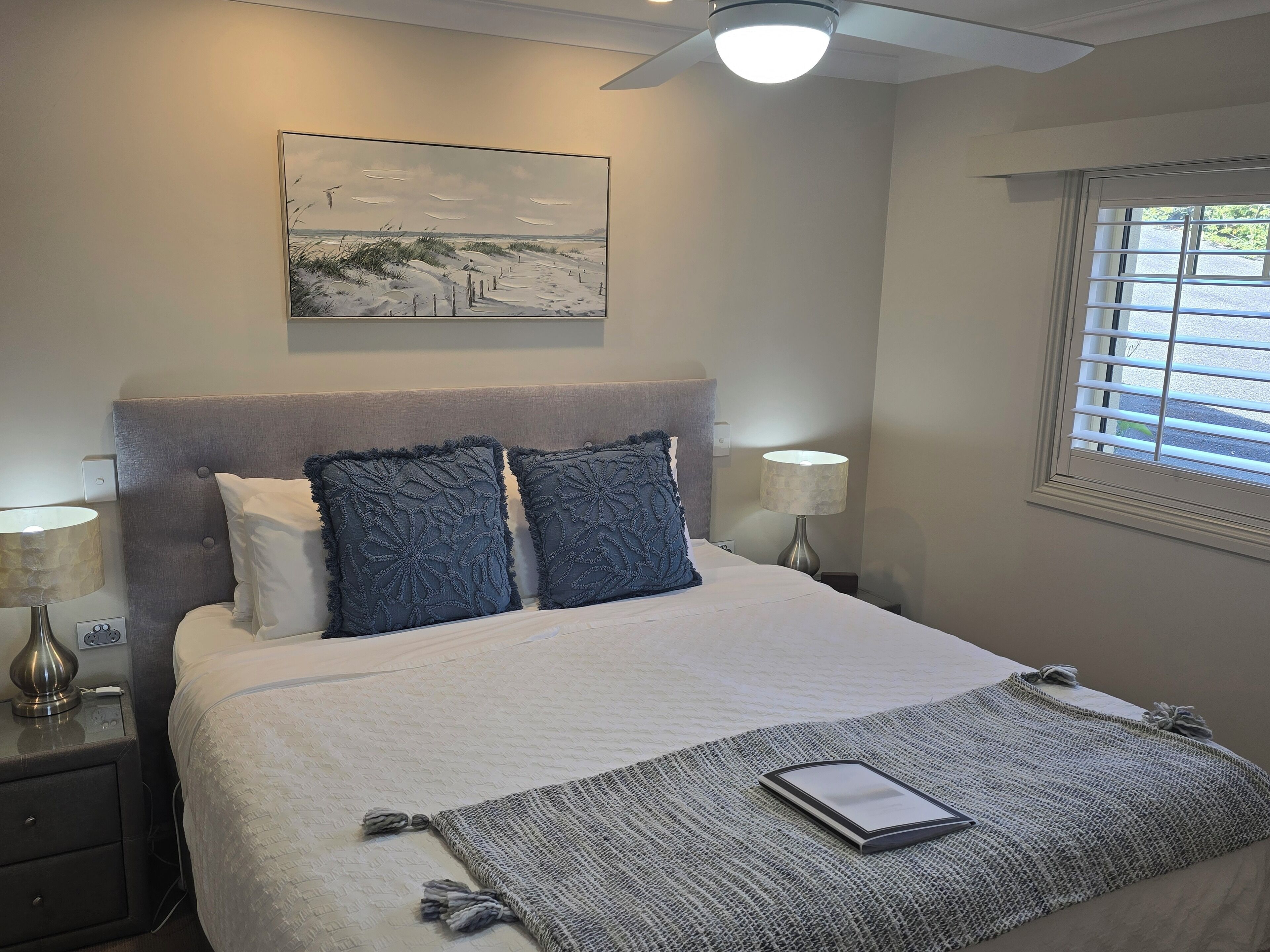 Ein ordentlich eingerichtetes Schlafzimmer mit einem großen Bett, zwei Nachttischlampen, einem Deckenventilator und einem Fenster mit Jalousien.