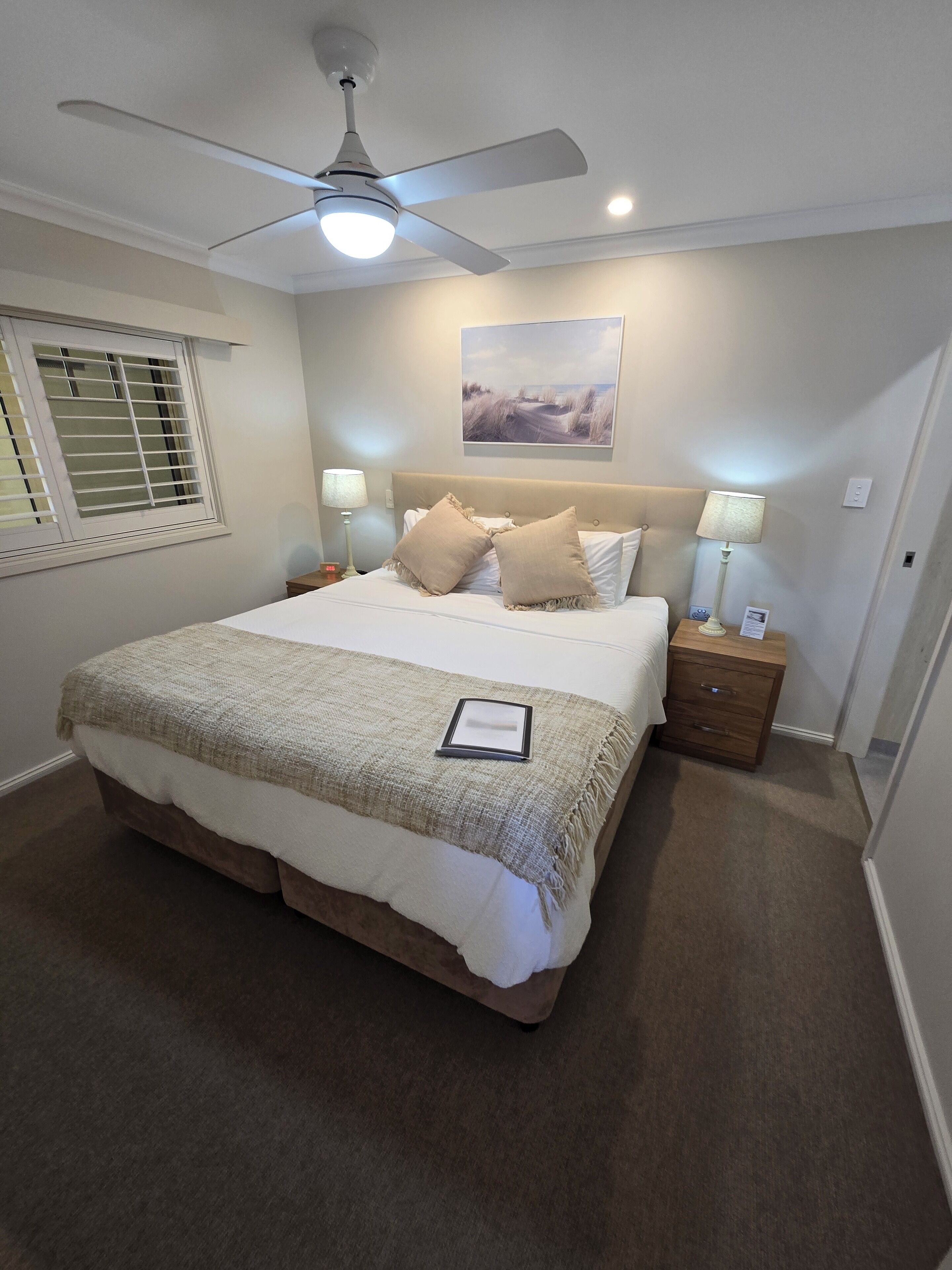A neatly arranged bedroom with a large bed, a ceiling fan, bedside lamps, and a window with blinds.