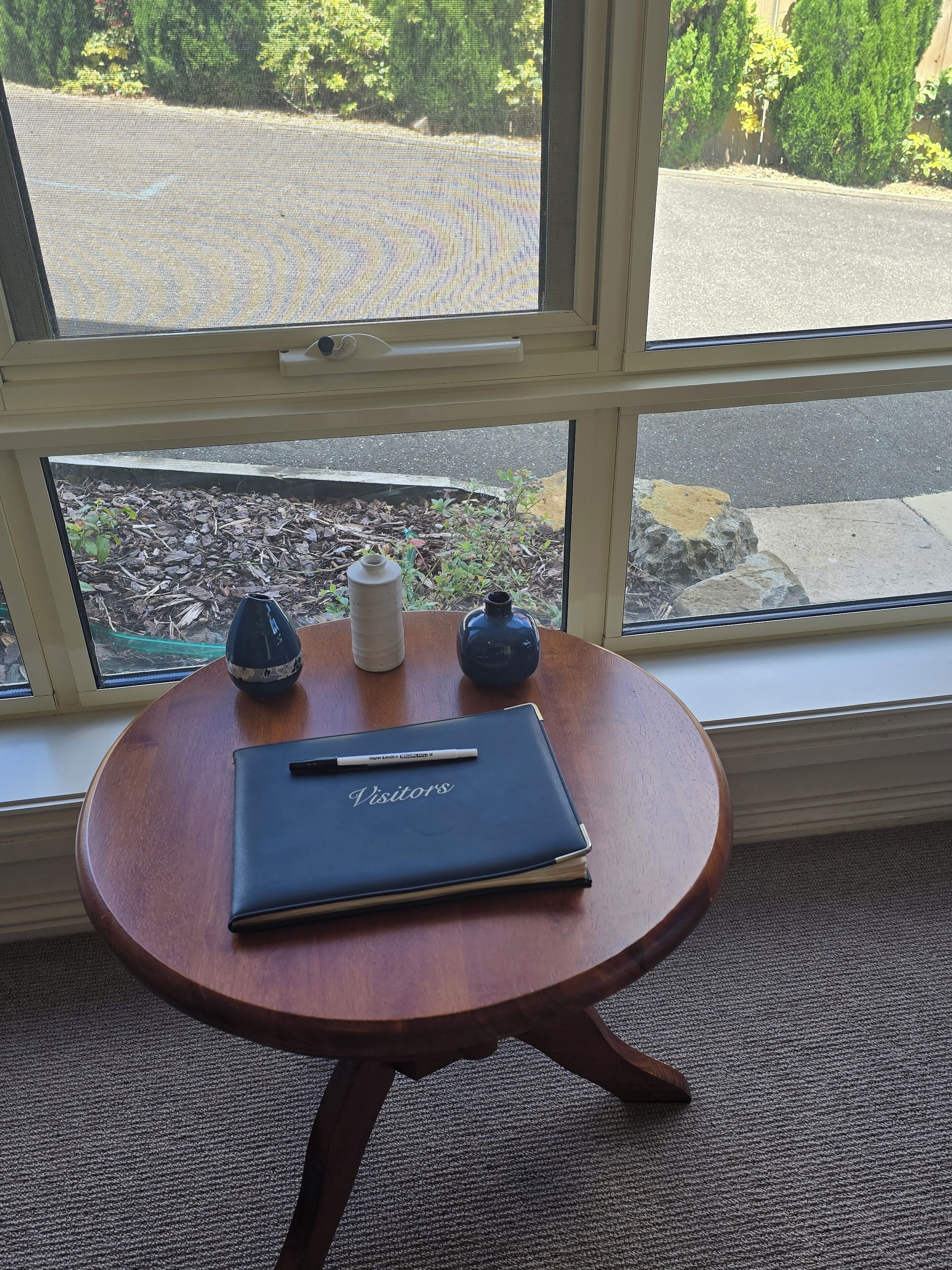 A small wooden table with a notebook labeled "Visitor's" and two bottles, positioned by a window with a view of a street and greenery.