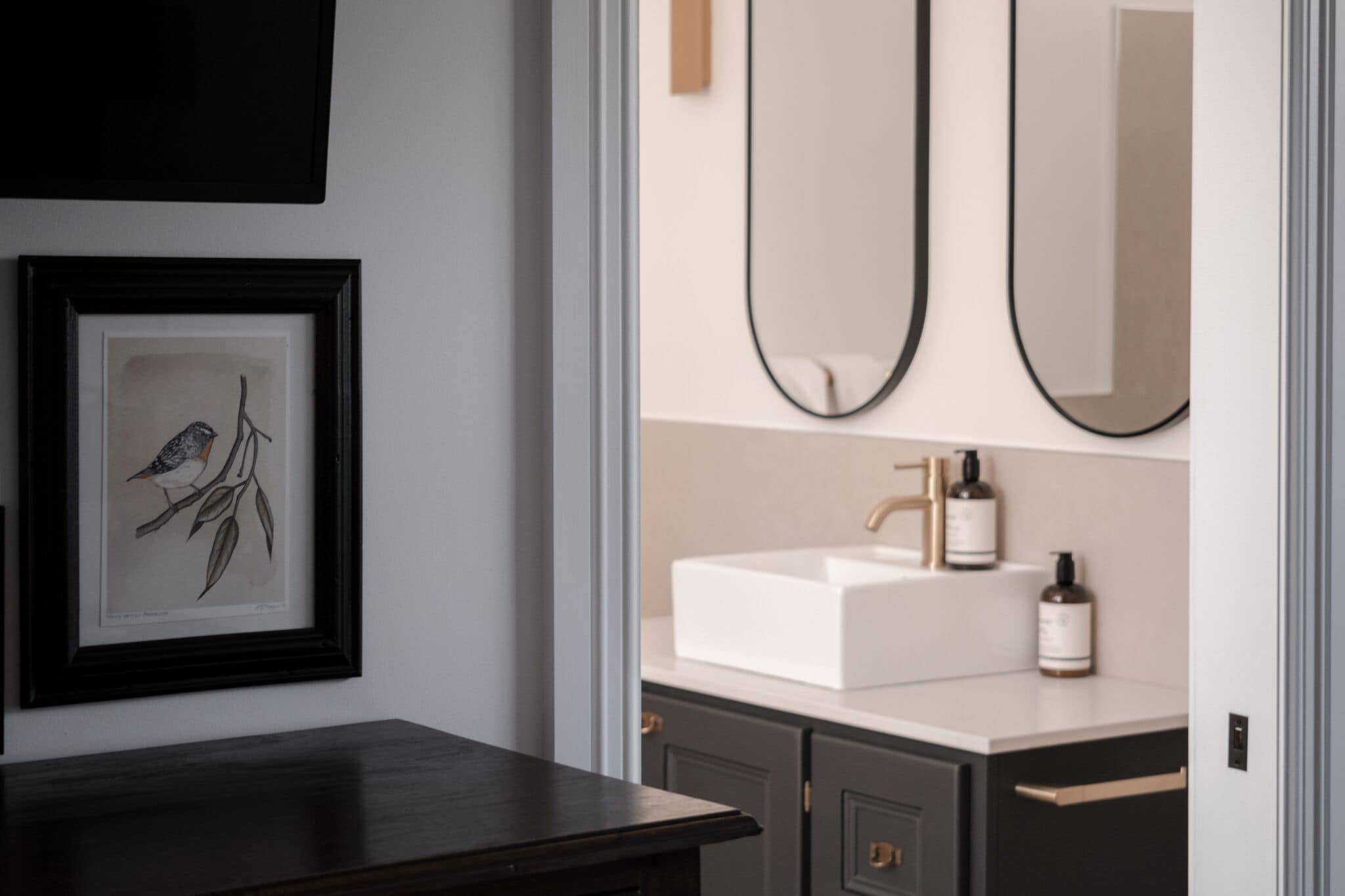 A bathroom with a white sink, two mirrors, and a framed artwork of a bird on a branch.