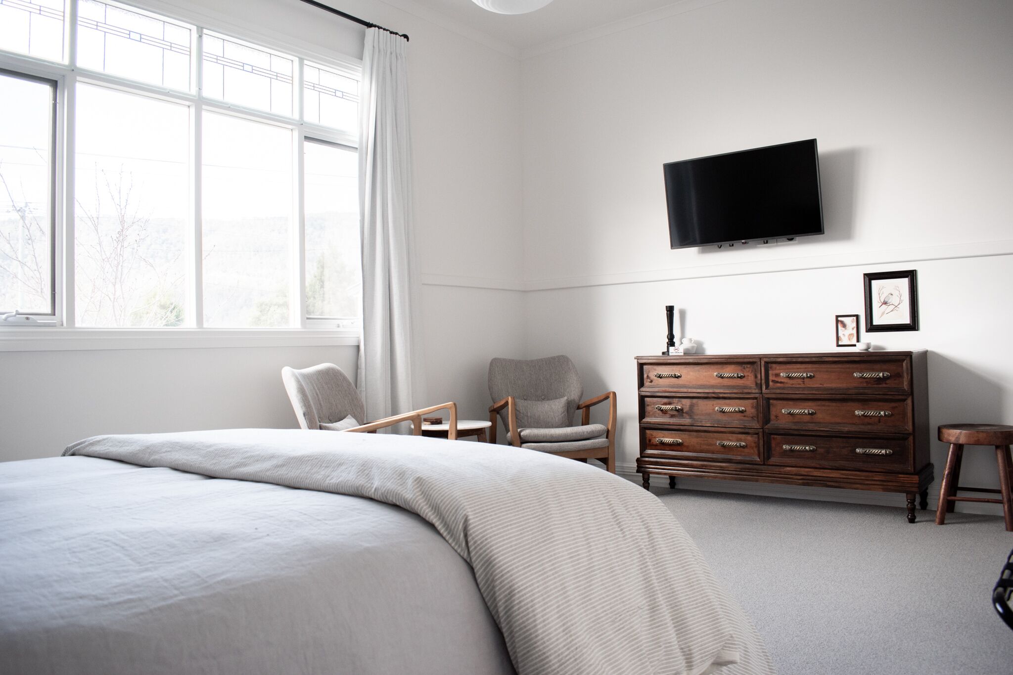 A bedroom with a large bed, a wooden dresser, a television mounted on the wall, and a window with curtains.