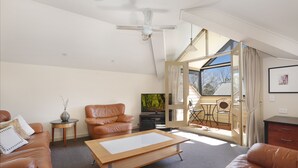 A modern living room with a brown leather sofa, a glass-top coffee table, a ceiling fan, and a large window with curtains. - Briars Country Lodge (Burradoo)