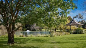 A gazebo with a conical roof, surrounded by lush green grass and trees, near a calm lake. - Briars Country Lodge (Burradoo)